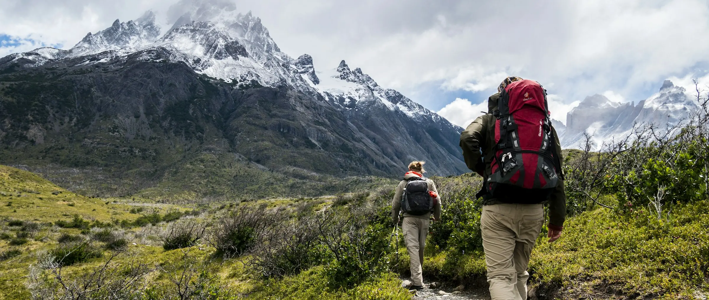 Two trekkers with backpacks and walking poles hiking through alpine landscape with snow-capped mountains